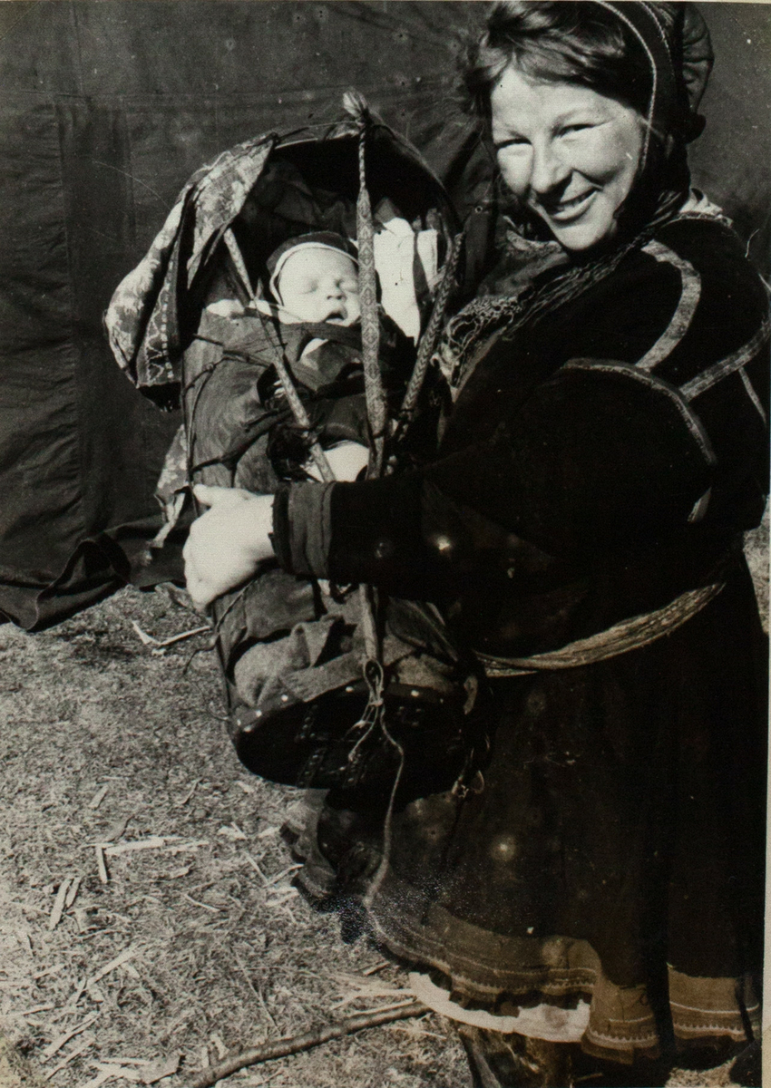 Bildet viser en samisk kvinne som kneler og holder gutten sin i en komse i sin fang. Barnet sover mens hans mor ser inn i kameraet med en stolt smil. Bak dem ser man et telt. 