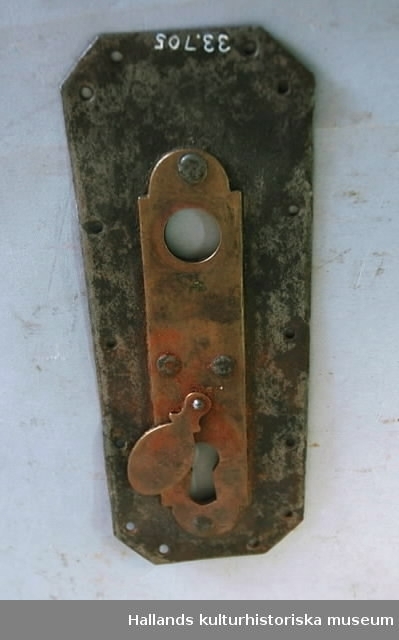 Beslag av mässing och järn. Hål för handtag och nyckel. En ledad bricka vid nyckelhålet.