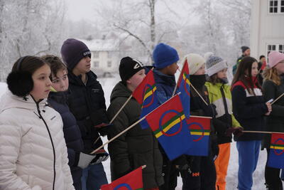 En gruppe 6.-klassinger står ute, foran Eidsvollsbygningen, og lytter til det som blir sagt. De er vinterkledd og holder samiske håndflagg.