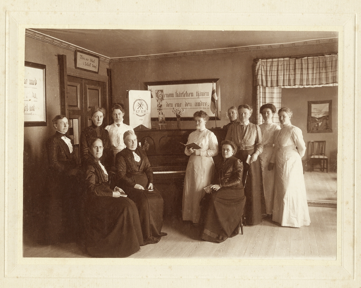 En grupp kvinnor i olika åldrar har samlats kring ett piano i en föreningslokal. I händerna har flera av dem något 
som ser ut som sångböcker. KFUK i Växjö 1912.