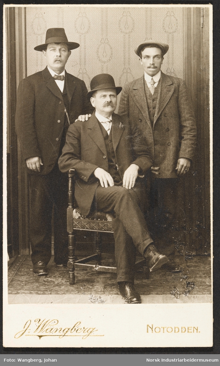 Visittkortfoto, tre menn fotografert i studio. Alle tre er kledd i dress. En av mennene sitter foran de andre i en stol.