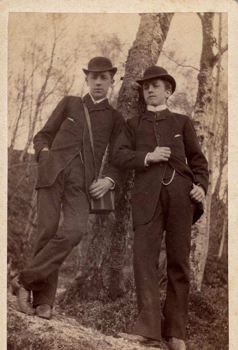 Portrettfoto. To unge menn i dress og bowlerhatt lener seg til et tre.