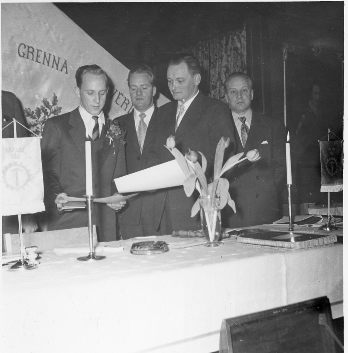 Fyra kostymklädda män med papper i händerna står bakom ett bord med tända ljus, tulpaner och bordsstandar. Bakom dem möjligen en fana med texten: "Grenna Hantverksförening"
