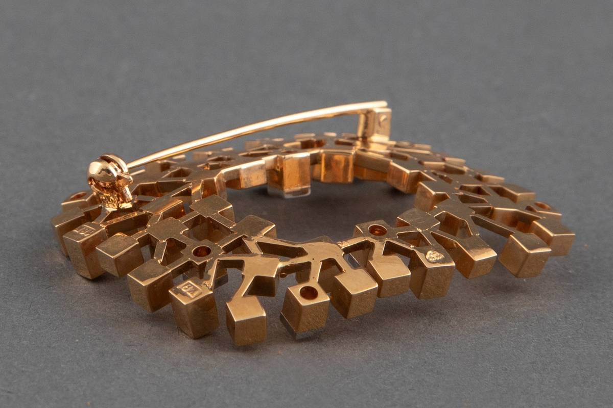 Brosje bygget opp av små kubiske elementer av vekslende lengde med endeflater som er cirka 3 x 3 mm. Brosjen er laget av 18 karat polert gull med 8 briljanter. Den er åpen i midten og har en nål på baksiden.