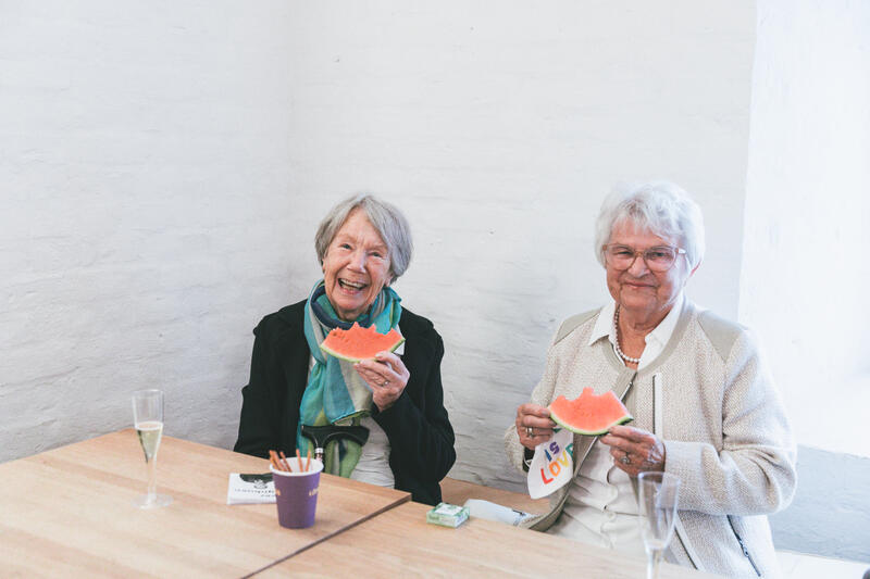 To eldre kvinner spiser melon i et rom med hvite murvegger. De smiler.