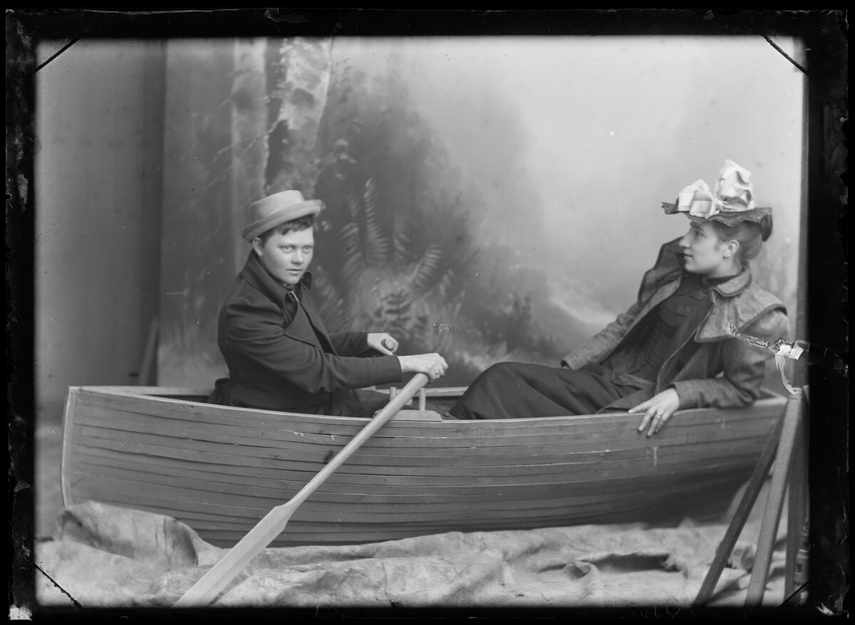 Two people in a rowing boat, placed in studio. One is dressed in a suit and straw hat, the other in a dress and hat with fabric flowers. The person in a suit, sits by the oars. Black and white photograph