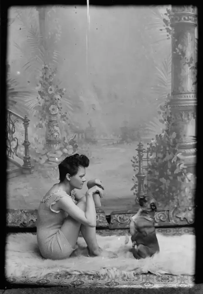 A woman with short hair and woolen underwear is sitting on a fur coat with a dog. The dog and the woman imitate each other's pose, with their paws and hands up in front of them. Black and white photograph.