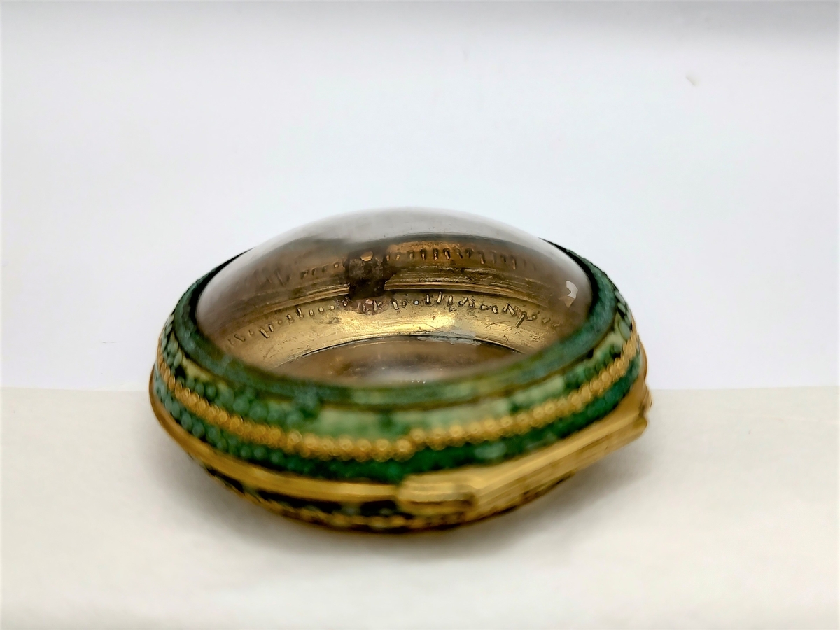 Sirkulær urkasse av forgylt metall, hengslet lokk med sirkulært glass i midten. Langs kanten er det pålimt grønn mosaikk med forgylt perlekant i midten.