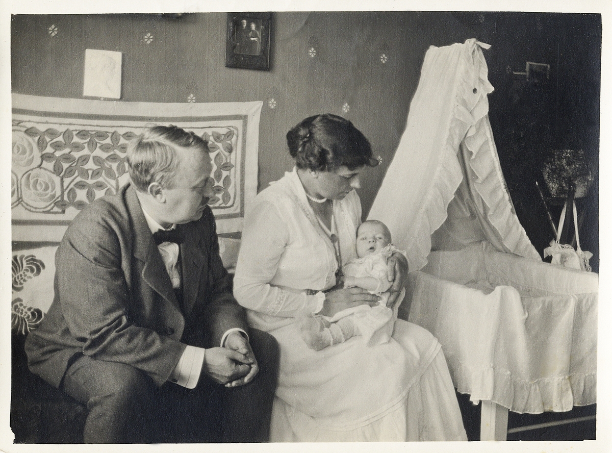 En man och en kvinna sitter med en baby i ett sovrum. Till höger syns en babysäng med
sänghimmel.