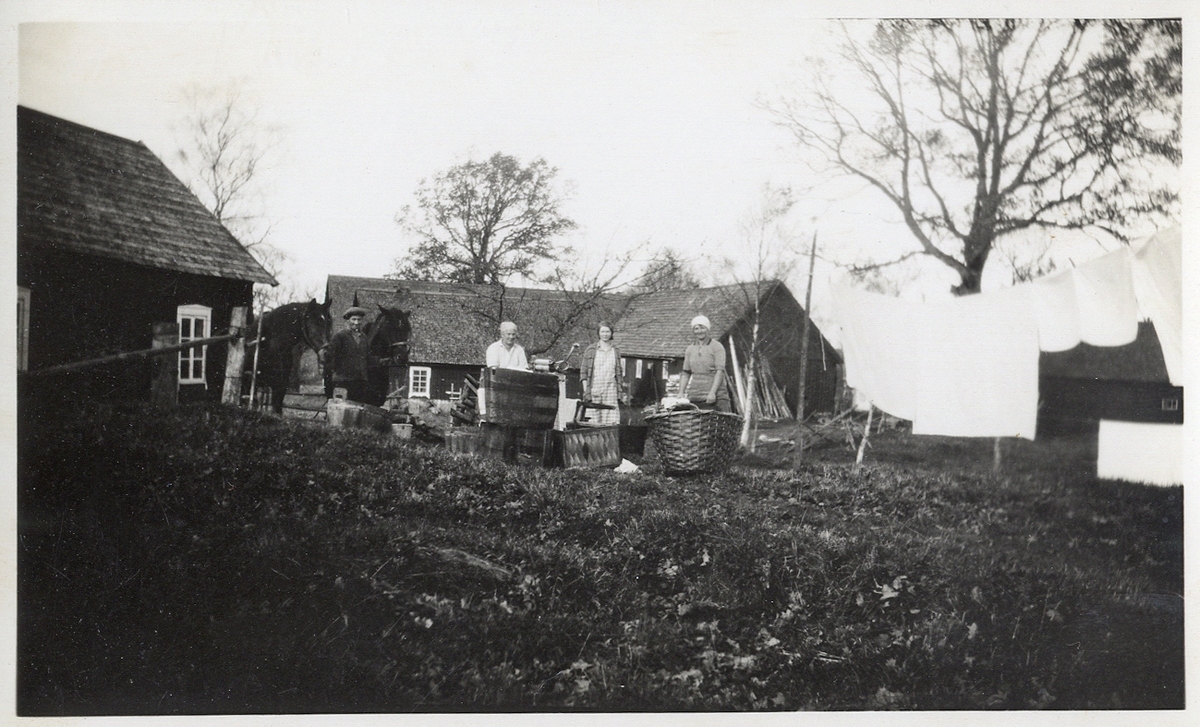 Bykning av tvätt utomhus vid en bondgård. Tre kvinnor är sysselsatta med storbyk. 
Till vänster syns en man i keps, med två hästar.