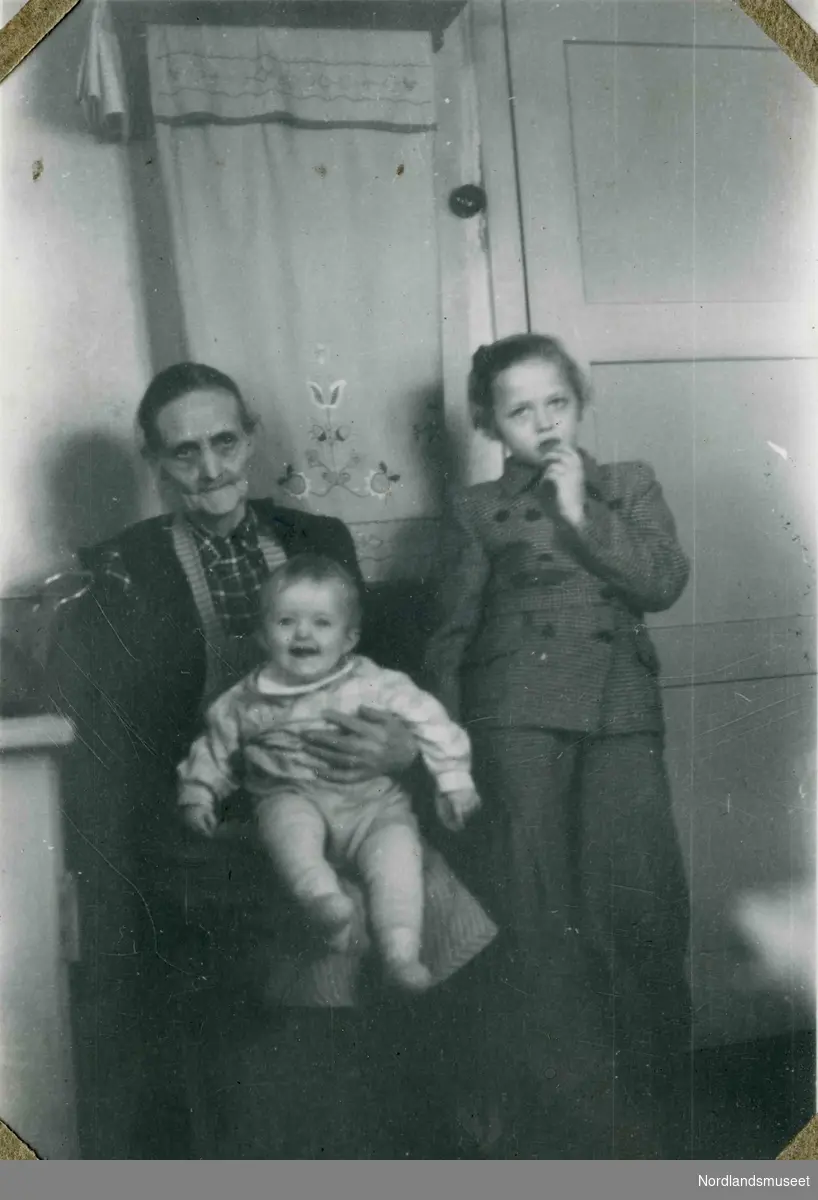 En eldre kvinne og to barn. Fotografert innendørs. Brodert pyntehåndkle i bakgrunnen. Tekst i album: Februar 1947.