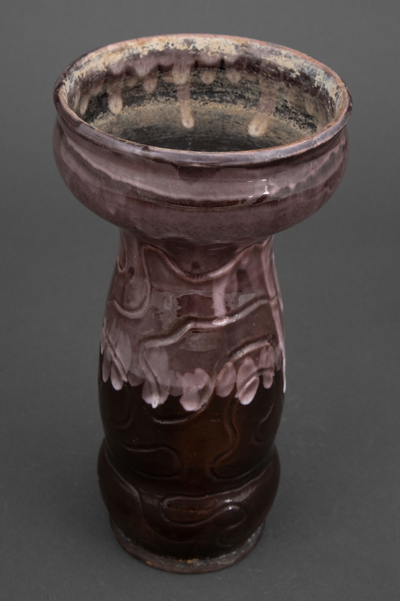 Vase i rødt gods med innrissede linjer som slynger seg rundt korpus. Nederste del av er glasert med blank glasur i brun, mens den øverste delen er dekket av brunfiolett rennende glasur med hvite partier.
