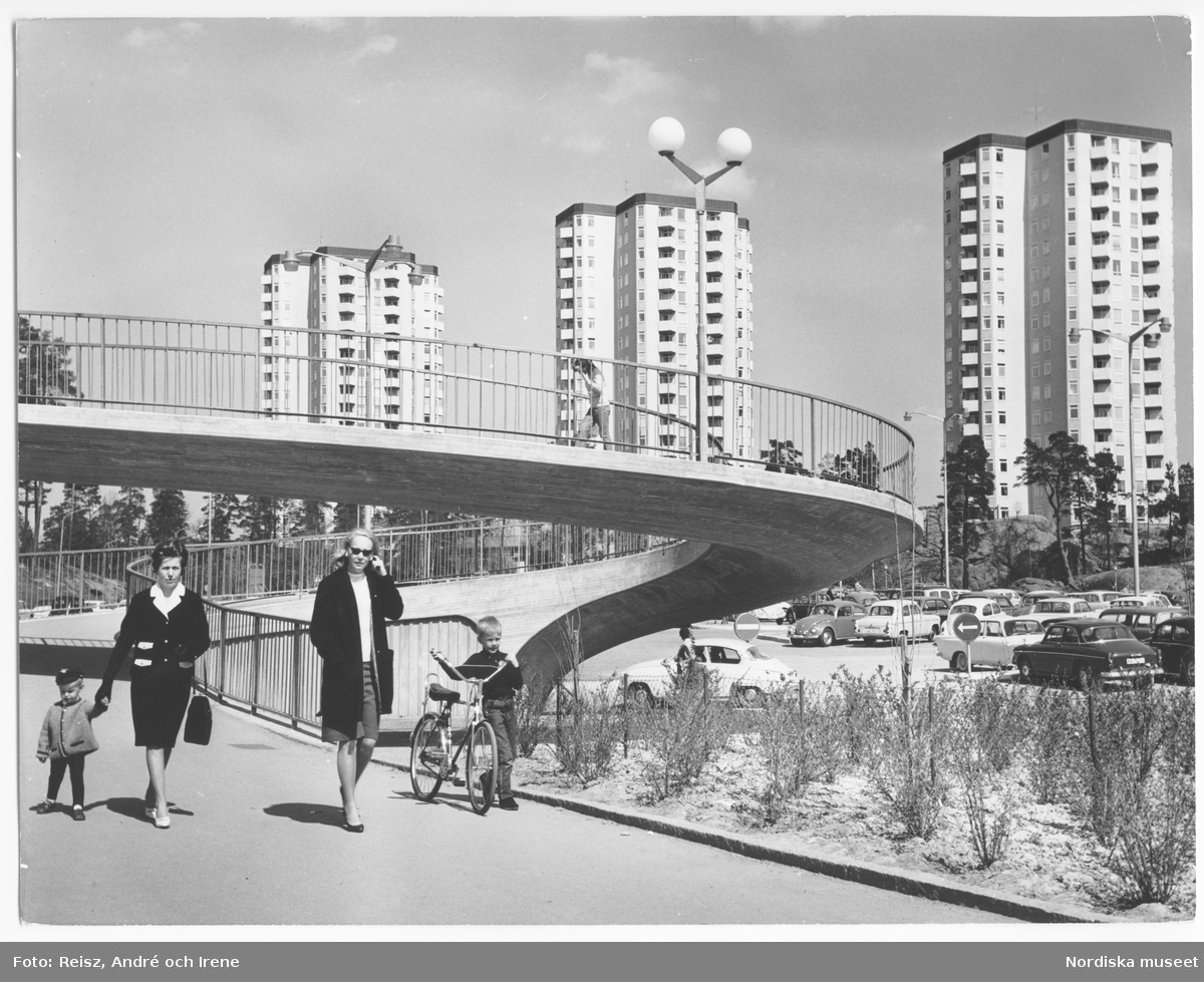 Stockholm. Farsta. Två kvinnor och två barn promenerar. Bakom dem en parkering och det nybyggda kvarteret Hjälmö, Farstavägen 87, 89, 91 med 17 våningar och 55 meter höga höghus,  byggda 1961 och ritade av Arkitekter Backström & Reinius.