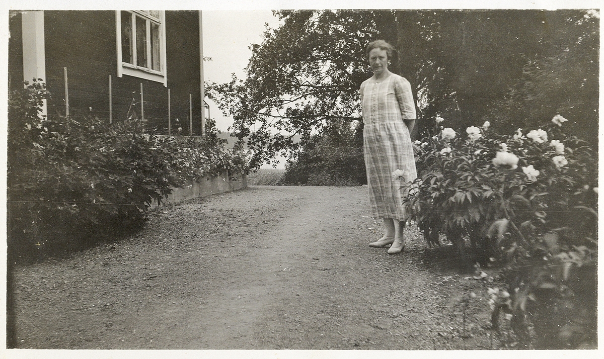 En kvinna i rutig sommarklänning poserar i en trädgård. Till vänster syns ett bostadshus.
Trol. i närheten av Sigtuna.