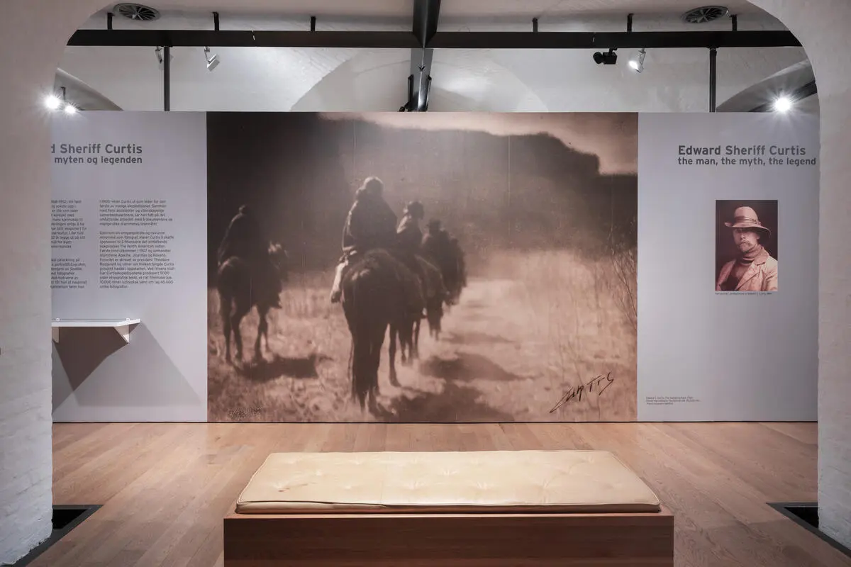 Photo from the exhibition Edward S. Curtis, the man, the myth and the legend. The picture shows one of the exhibition rooms, on the wall there is a large picture of some indigenous people riding across a landscape with mountains around.