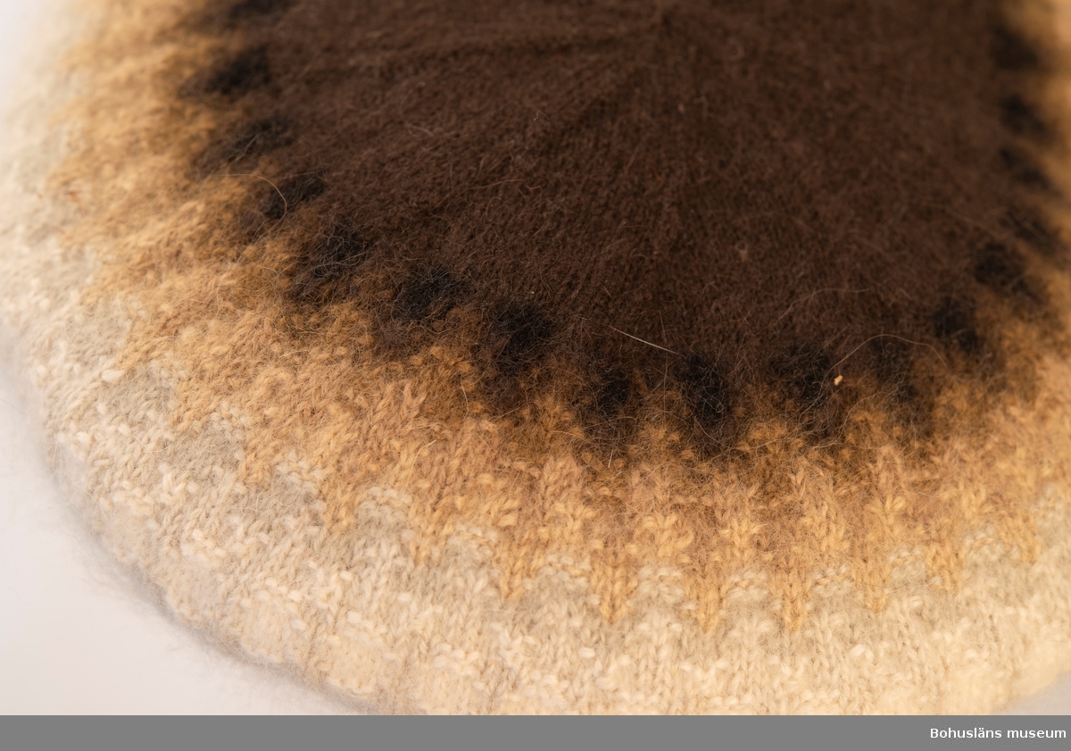 Stickad barett med slätstickade enfärgade partier med en resårstickad kant. Baretten är mönsterstickat med räta och aviga maskor i mörkbrunt, svart, ljusbrunt, brunbeige, grått och vitt mot en brun botten.
