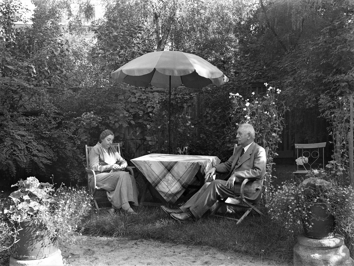 Borgmästare Johan H Linnemann Janssen sitter tillsammans med frun Annie (f Ullgren) i sin lummiga trädgård under ett parasoll i kv Komedianten 1; ett av de s k Tvillinghusen. Annie handarbetar. Spaljerade luktärter blomstrar i bakgrunden och pelargoner står i urnorna vid sandgången.