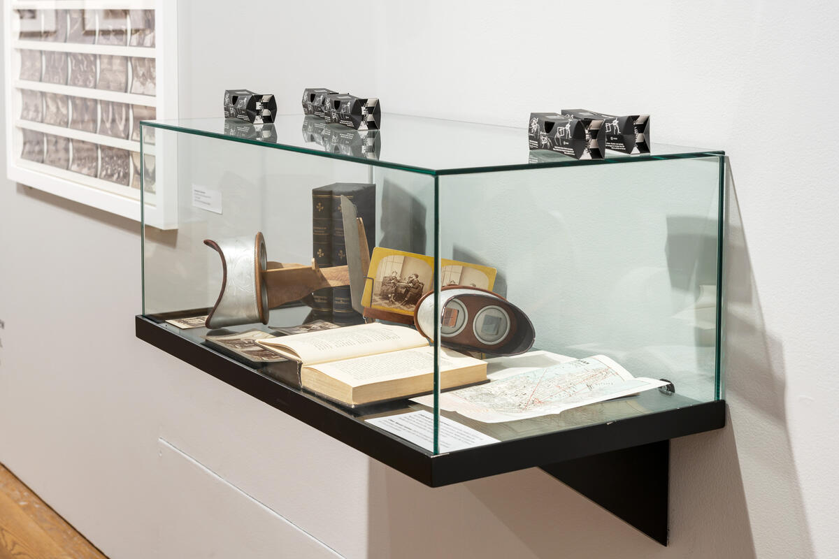 Documentation photo from the exhibition World in 3D. The picture shows a section of an exhibition room with a stand with stereo speakers and stereo photographs. In addition, you see yellow stereo monitors mounted on the wall where the public can look in and see 3D images. There are small stairs below which ensure that children can also see.