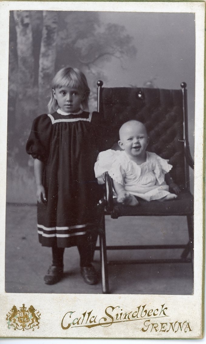 Kabinettsfotografi: ett skrattande spädbarn sitter i en stol. En flicka i mörk klänning med ljus bård står intill.
