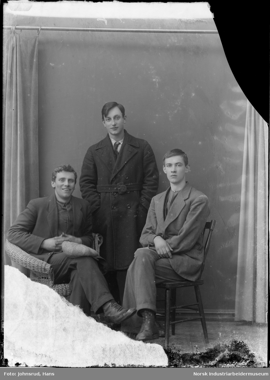 Portrett av tre menn, to sittende i dress og en stående med frakk mellom dem.