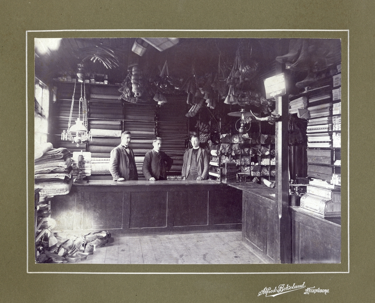 Tre män står bakom affärsdisken i en s.k. manufaktur- och diverseaffär, trol i Helsingborg. På hyllorna trängs bl a en hel del tygrullar m.m.
Mannen till vänster ev. Carl Ljunggren (1873-1956), som var verksam i Helsingborg vid denna tid.