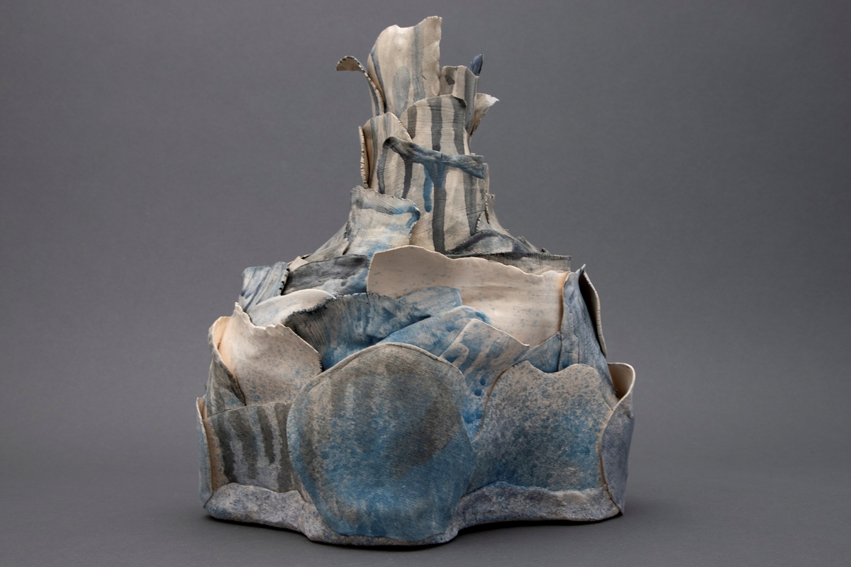Skulptur/ objekt i abstrahert vaseform. Bygd opp av flere lag med tynne modellerte flak av papirleire, porselen og steingods. Farget med koboltblå farge og klar glasur.