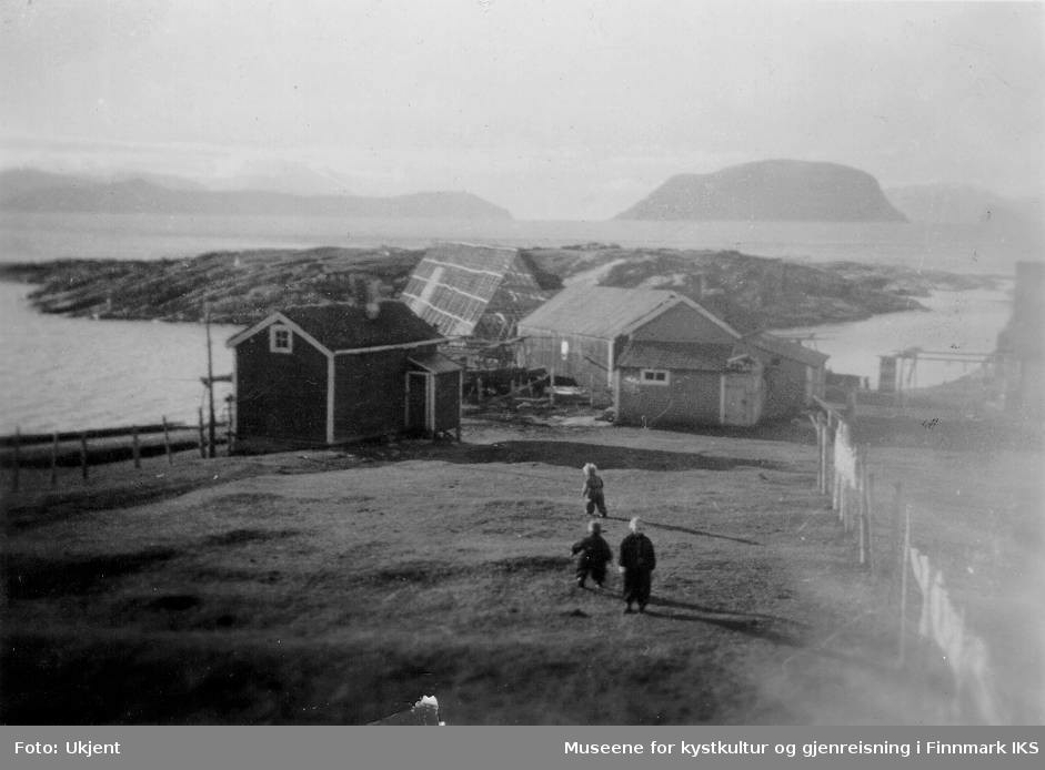 Tre barn står på en eng på Melkøya i Finnmark. Bak dem ser man flere bygninger, et fiskehjell og et svaberg. Langt i bakgrunnen ser man Seiland, Håja fjellet og Sørøya bak Sørøysundet.