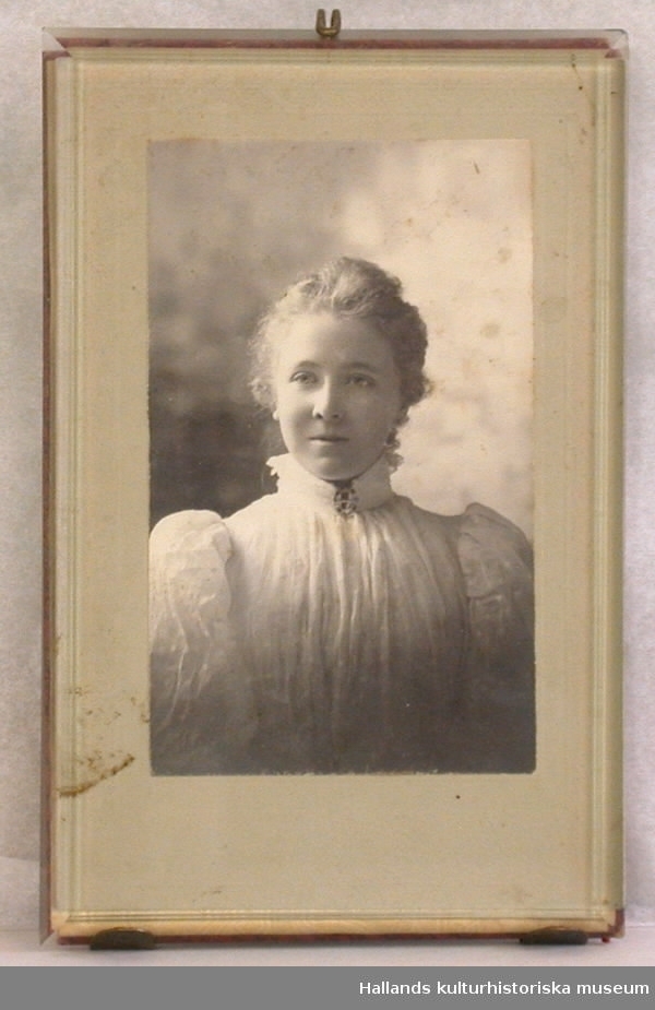 Fotografi. Rektangulärt. Med grå passepartout. Föreställer Alice Bexell från hennes Amerikatid på 1890-talet. Glasat. Metallställning på baksidan för att kunna ställas på ett bord eller dylikt.