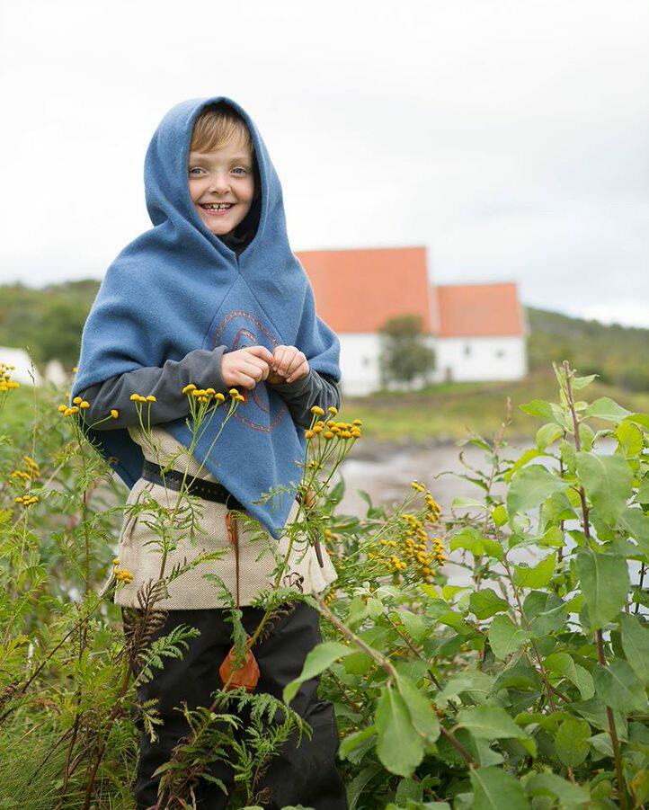 En gutt i vikingklær omgitt av blomster. En hvit kirkebygning med rødt tak i bakgrunnen.