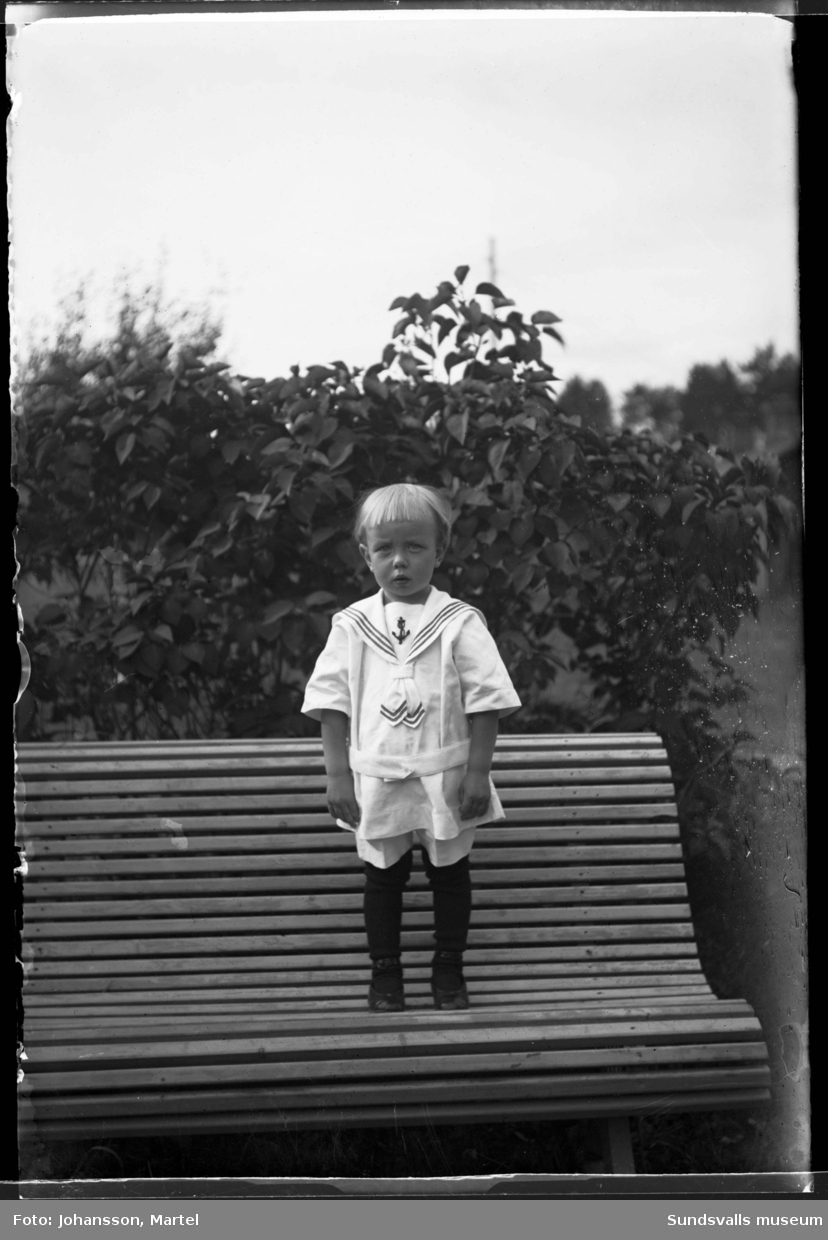 Porträttbild av en liten pojke i sjömanskostym, han står på en soffa ute i trädgården.