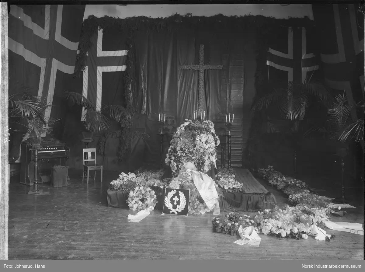 Begravelse til skolebestyrer Evje. Kiste omringet av blomster og blomsterkranser med tekstsløyfer. Norske flagg henger på veggene.