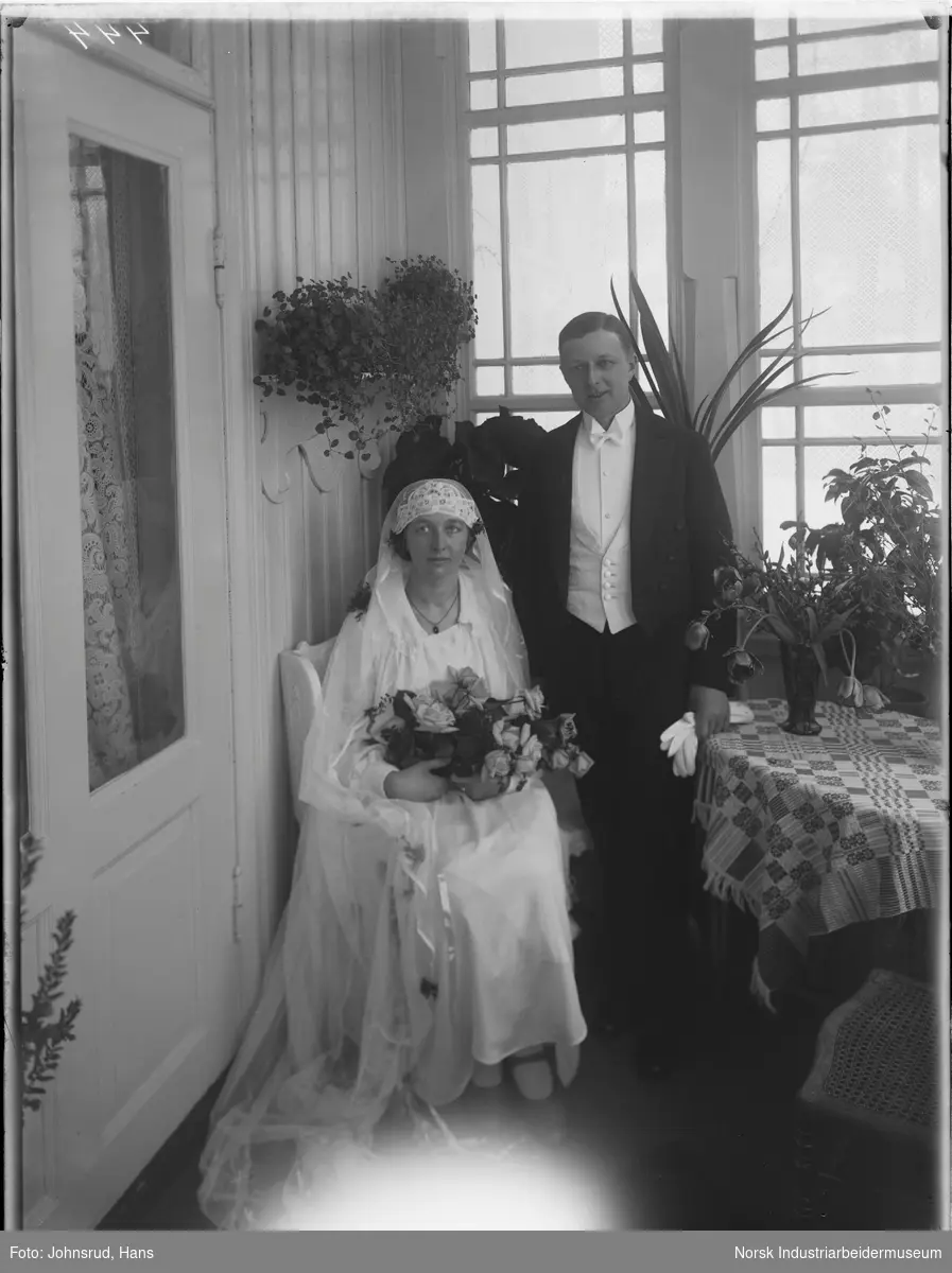 Bryllupsbilde av brudepar oppstilt innendørs i overbygget verandabygg. Bruden har slør på hodet og brudebukett på fanget, brudgommen er kledd i kjole og hvitt.