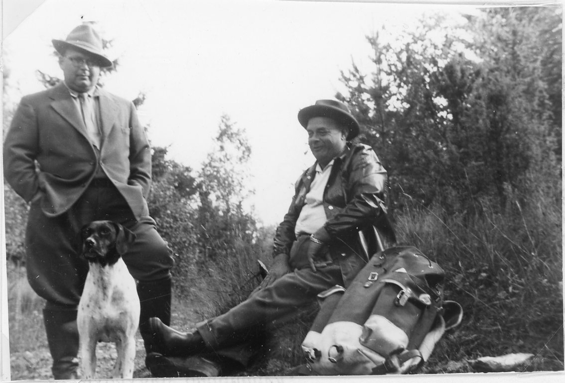 Troligen två jägare som tar en paus vid ett buskage. En man står med en hund framför sig, den andre sitter ner med en ryggsäck intill sig.