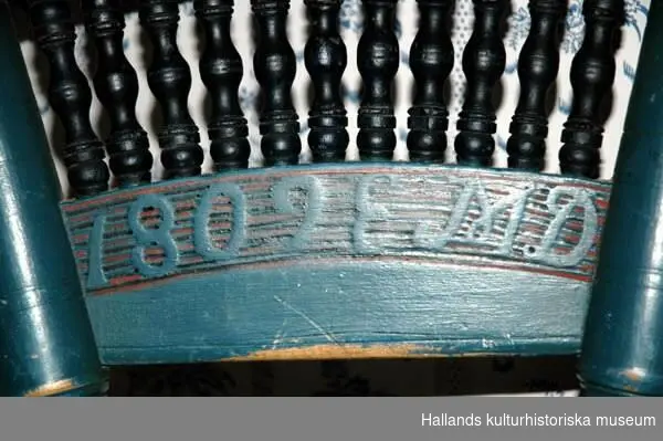 Stol av blåmålat trä. Tre snedställda, runda, profilerade ben. Rund sits, ryggstöd bredare uppåt, utsirad ryggtavla målad i rött, vitt och svart. Årtalet 1809 och initialerna E M D målade i blått på röd botten.
