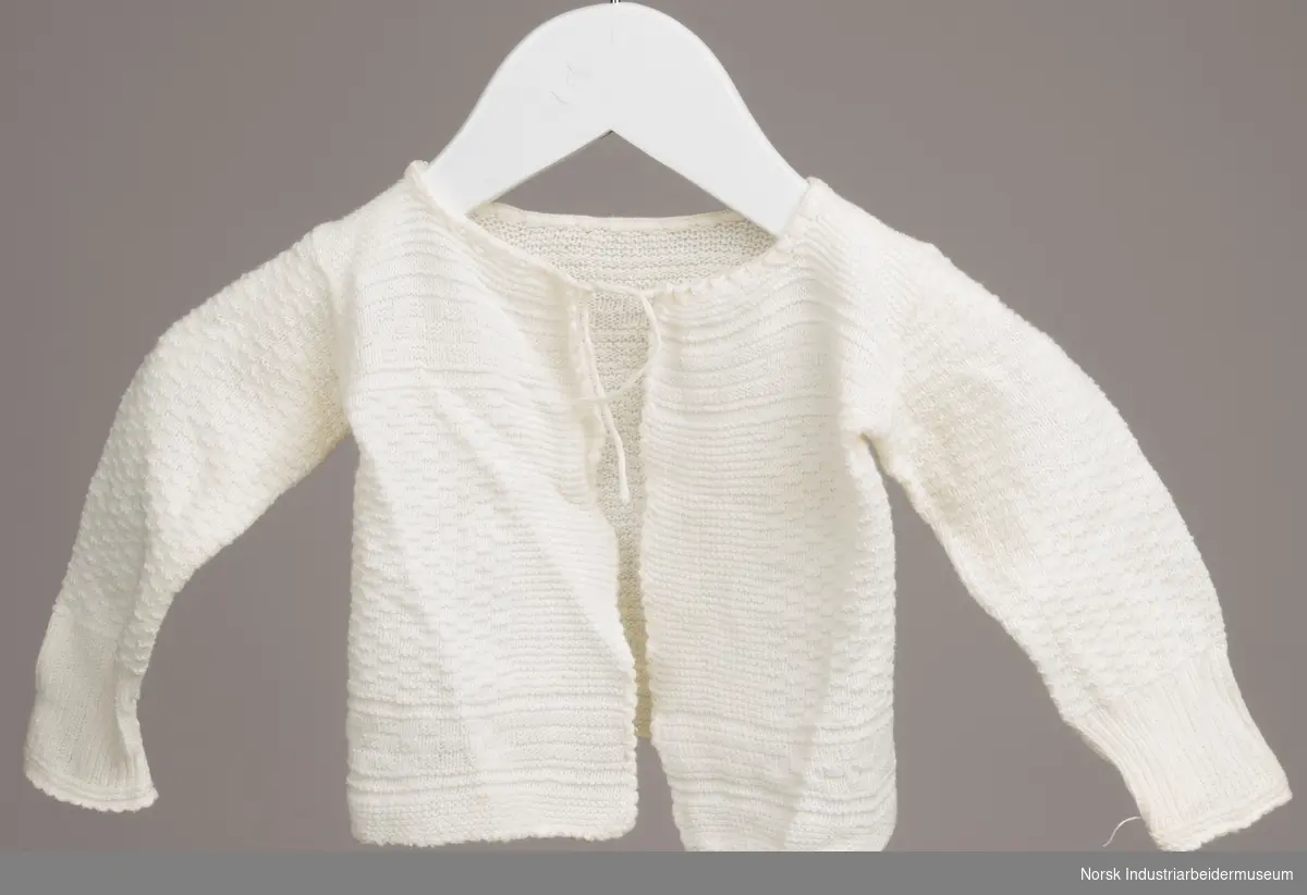 Hvit trøye til spedbarn med snor i halsen. Trøyen er maskinstrikket med mønsterstrikk i samme farge. Masker er løse i venstre armhule.