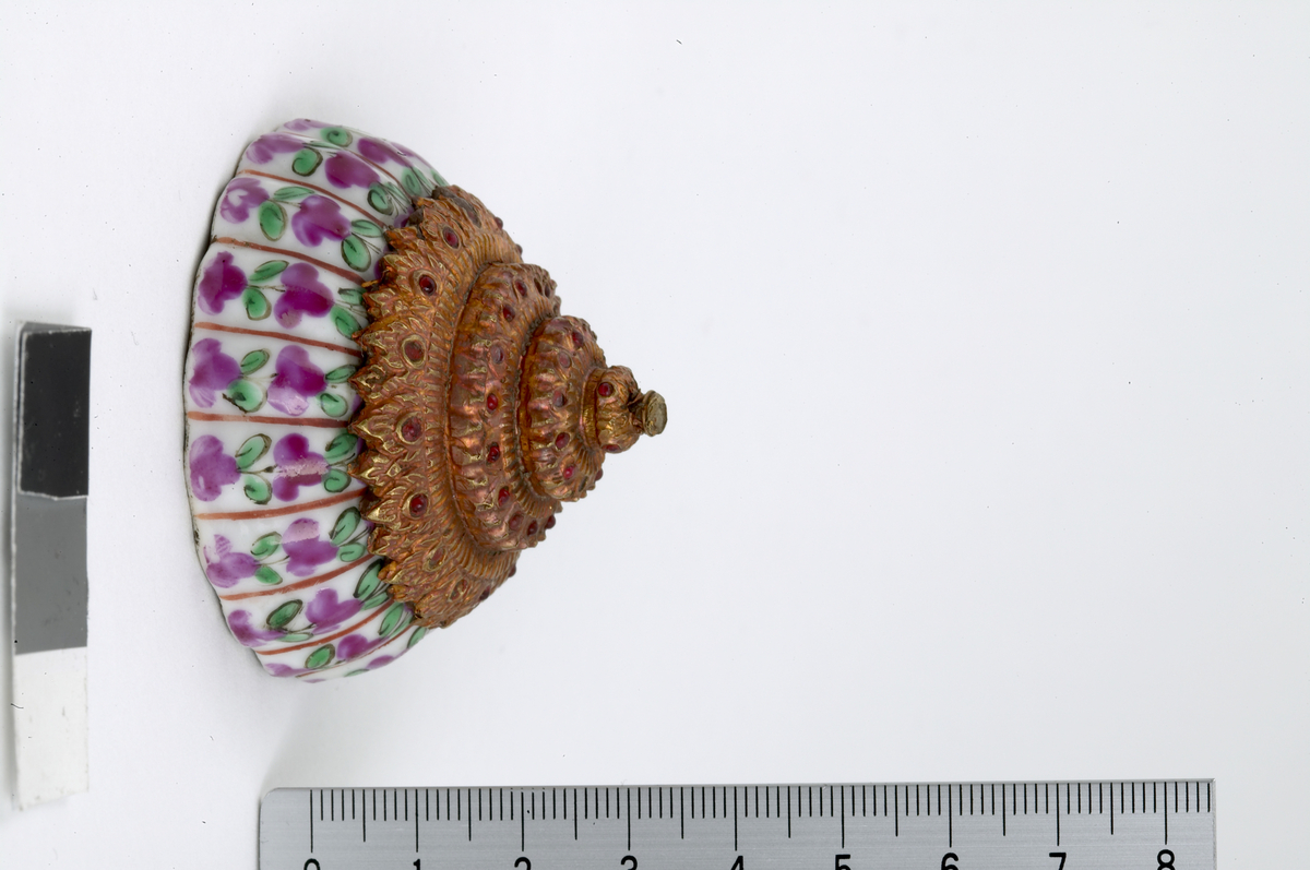 Bencharong. Subgroup Lai Nam Thong. Small cosmetic box with cover. The cover is in the shape of a stupa, covered with gold foil studded with glass gems. Ribbed body with vertical bands with small purple flowers and green leaves on white background.  The ribs are separated by red lines. Green footring. Cover height 4.2 cm, diameter 5.6 cm.19th century. 