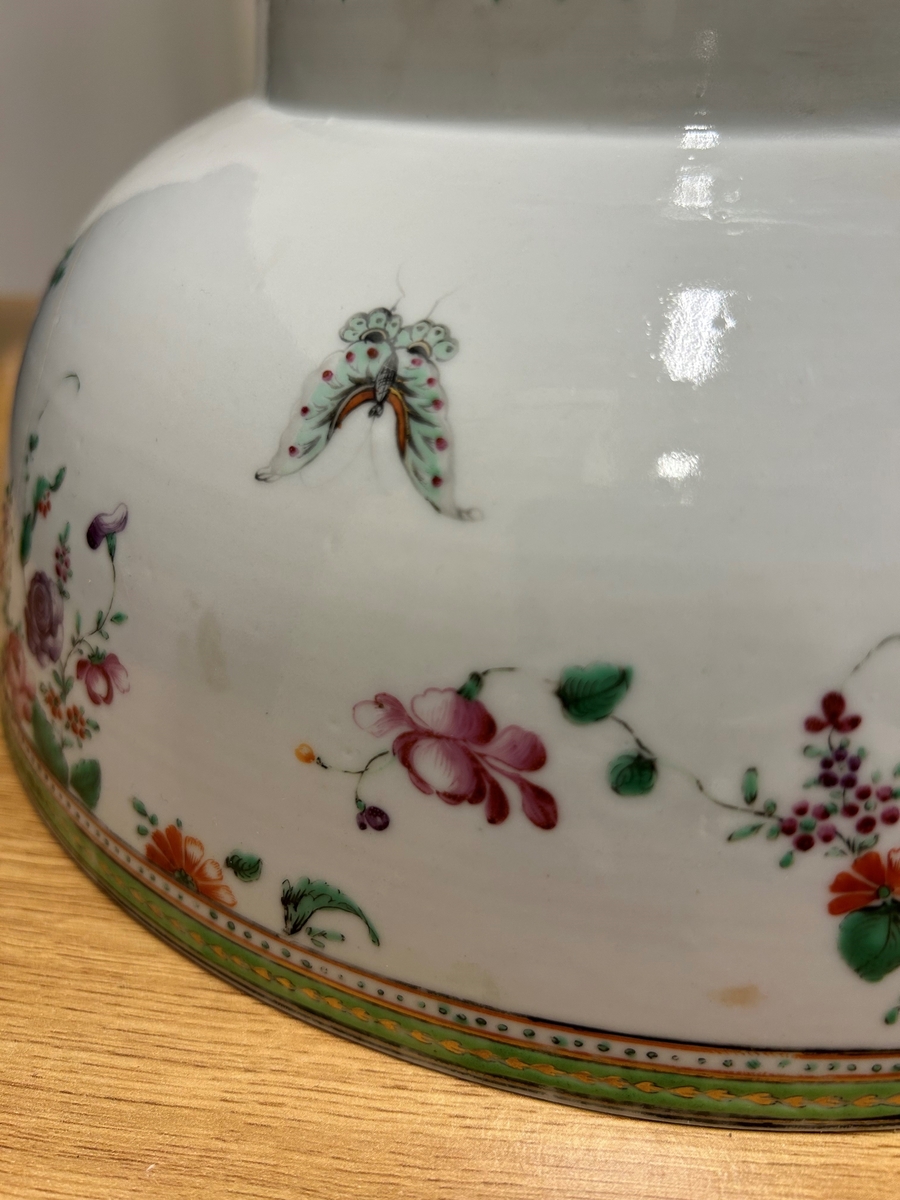 Vit cirkelformad skål av ostindiskt kompaniporslin med blommor i grönt, guld och rött, två fjärilar, en husfluga och en skalbagge. En markerad målad kant upptill utan och innanför på skålen. En blomma på botten av bålskålen. 1700-tal.