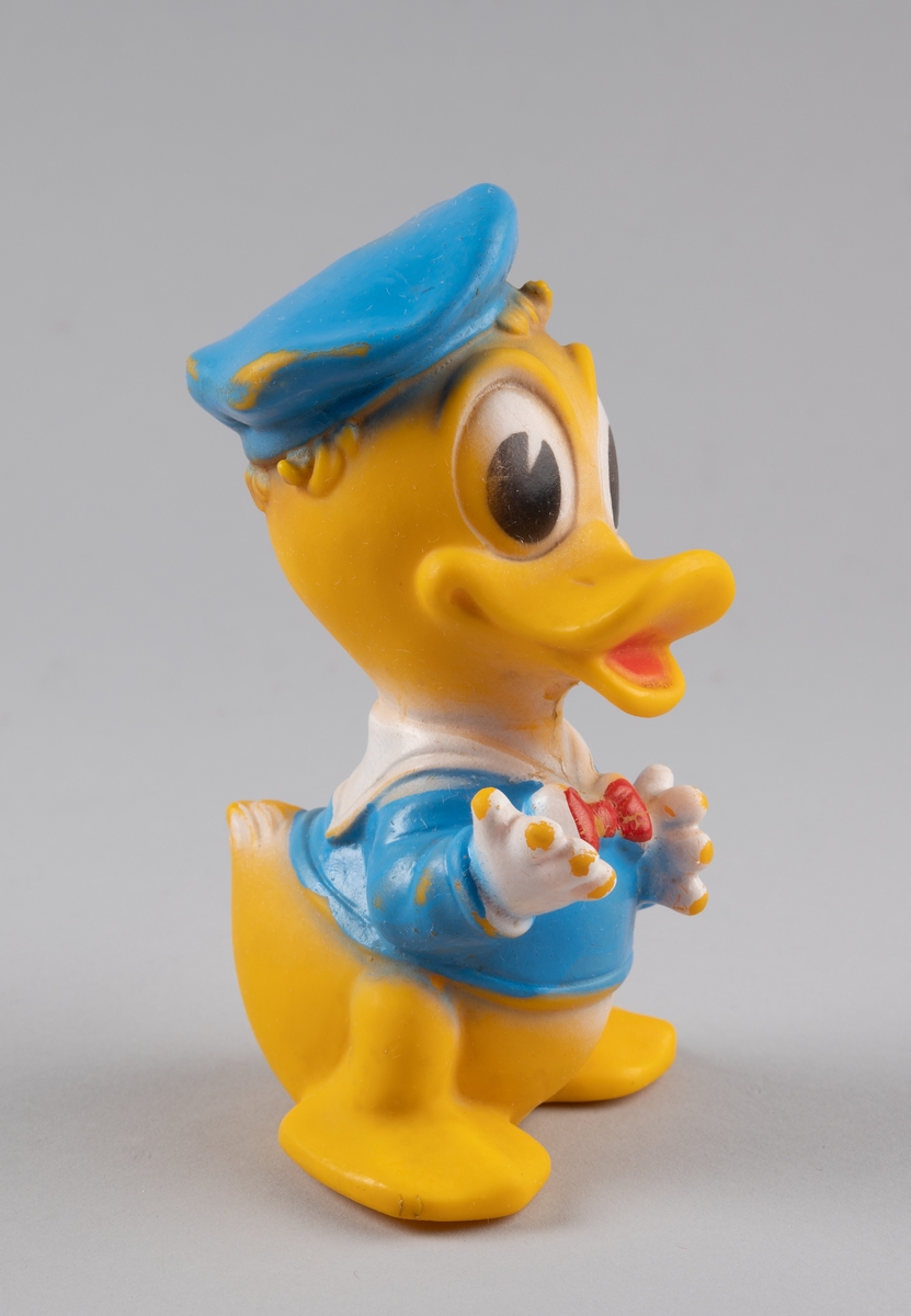Leketøy av mykplast i form av en Donald Duck. Den er gul med blå jakke med rød sløyfe, den har blå hatt og hvite hansker. Øynene er store og ovale, hvite og svarte.