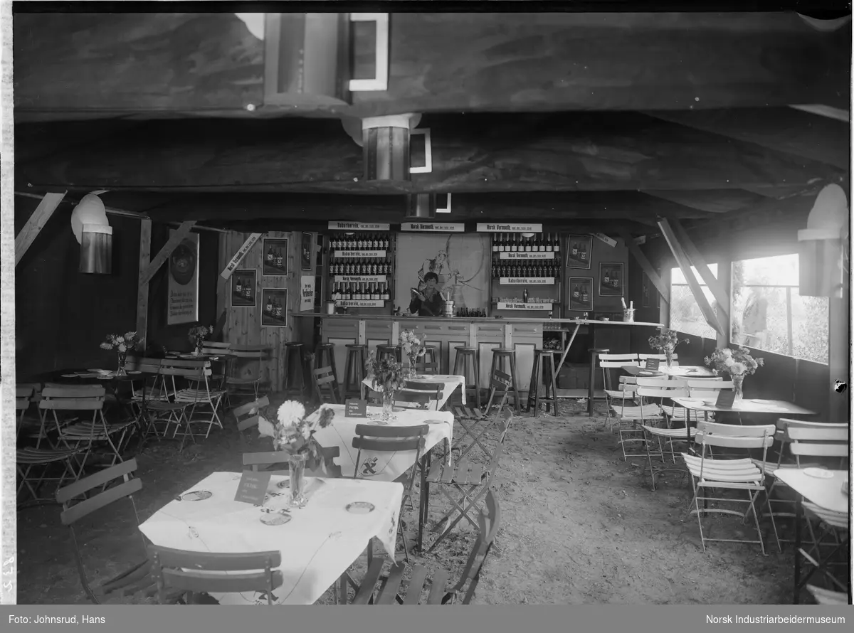 Handelsstevnet i Notodden 1933. Interiør for resturant og bar. Kvinne stående bak disken. Reklame hengende rundt i lokalet. Mann stående på utsiden av gjerde og ser inn.