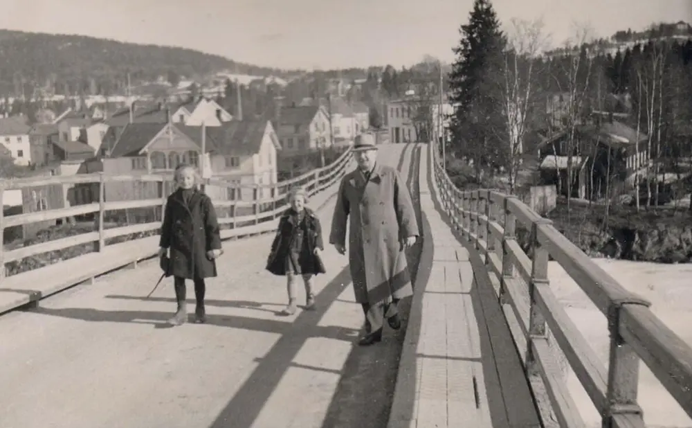 Svart-hvitt foto av Kongsvinger bru. En mann med hatt og frakk og to små jenter går på brua.