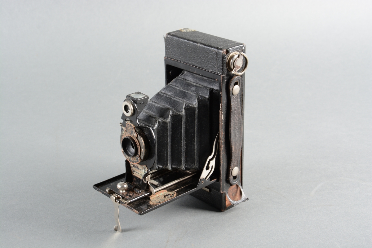 Foldekamera med futteral. Kameraet er svart og består av et rektangulært etui trukket med skinn. Den ene langsiden av etuiet har et skinnhåndtak, og på baksiden er et lite, rundt og rødt vindu. Inni etuiet er et rom for oppbevaring av film med en spole av plast. Selve foldekameraet, når det er foldet, er plassert inni etuiet som del av boksen. Når kameraet er foldet er det en rektangulær boks som på baksiden har en relativt stor linse med svarte tekstiler i konsentriske lag  (innsiden av foldene) rundt linsen. Kameraet kan tas helt ut av etuiet, og det kan åpnes ved å dra i et lite, nedslått metallhåndtak. Lokket er hengslet og kan låses i nittigraders vinkel slik at håndtaket blir en fot som kameraet står på. Kameraet kan dras ut og inn på skinner. Det har ulike skiver for justeringer.