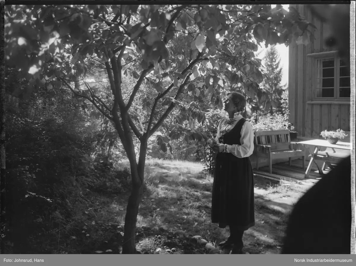 Kvinne i bunad stående under tre i hage. Bolighus med veranda og verandamøbler i bakgrunnen.