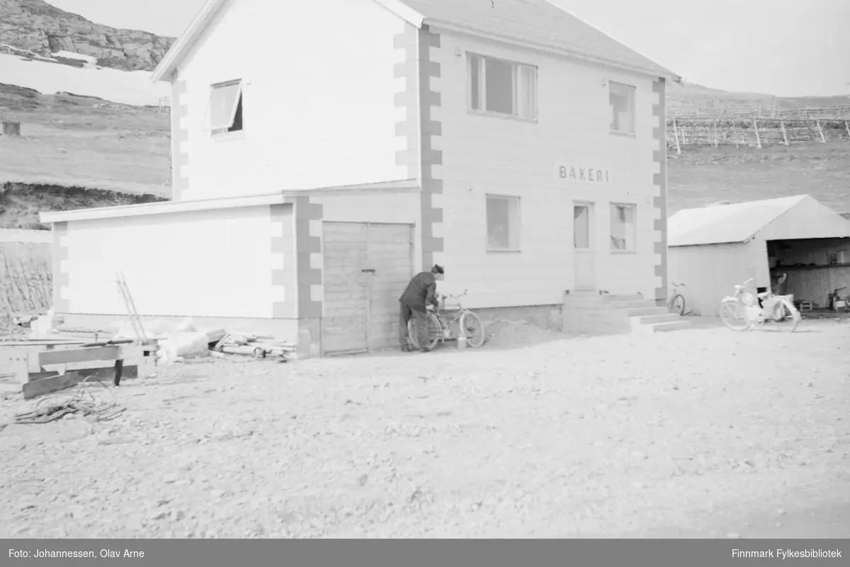 Foto av gateparti, kan være Ernst Eriksens bakeri i Fjordvegen i Båtsfjord (usikker identifisering)

I følge informant pleide barna i Båtsfjord å få kakeskrell gratis fra bakeriet på slutten av 1960-tallet

I midten kan man se et bakeri med en motorsykkel parkert utenfor