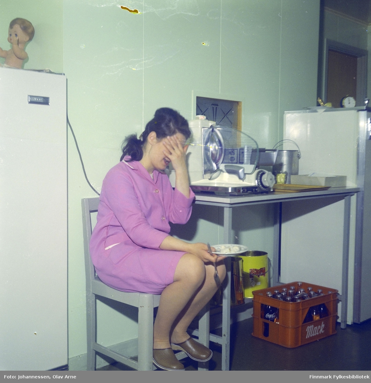 En ukjent kvinne spiser men hun sitter på en stol i et kjøkken, antagelig på Skansen i Båtsfjord

En kasse kan bli sett til høyresom inneholder coca-cola flasker

Foto trolig tatt på 1960-tallet