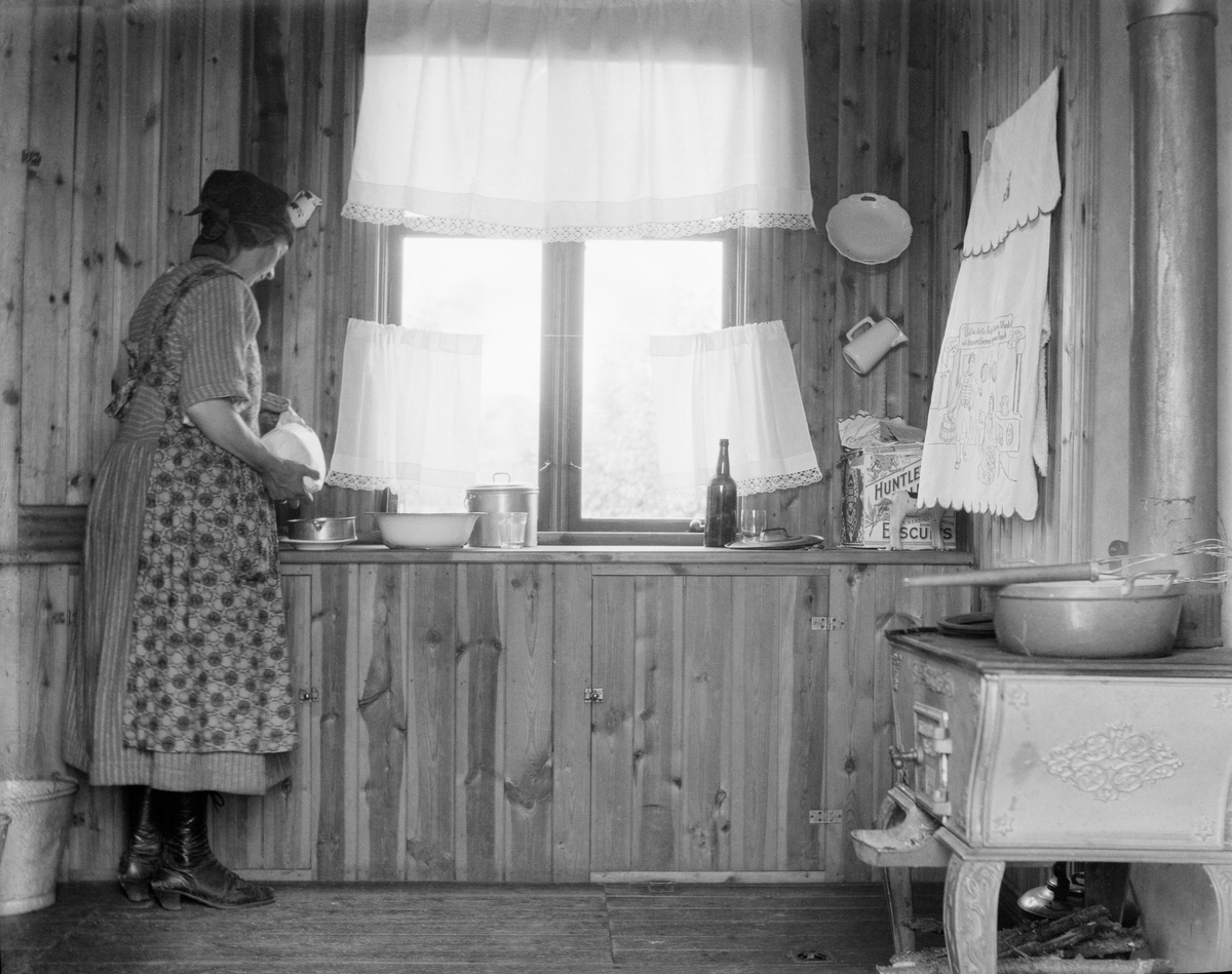 Bilde av ei kvinne, Anne Midttun, som ikledd forkle og skaut lager mat på kjøkkenet til Telemark planteskole, Det norske skogselskaps planteskole ved Gvarv. Hun står ved kjøkkenbenken og er i ferd med å helle over i ei lita form.