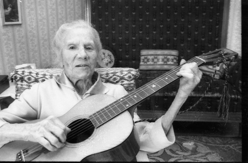 En äldre kvinna sitter i en fåtölj och spelar gitarr.