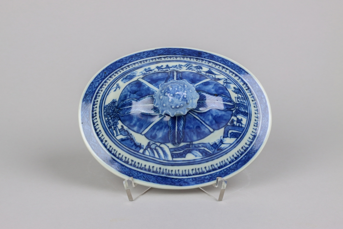 Oval shaped and slightly domed with knob in form of a flower head. On the top of lotus leaves and a list of pagoda lanscapes. The edge of the lid decorated with a dark blue  border in a criss cross pattern. All decor in blue underglaze.