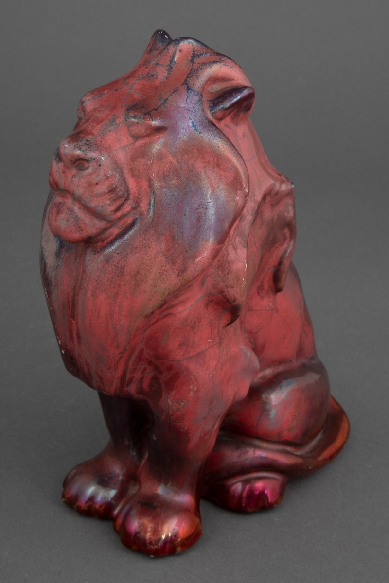 Keramikkfigur som viser en sittende løve. Løven er i lett, lysegrått gods. Figuren er glasert med mørkerød blank glasur og metall-luster.