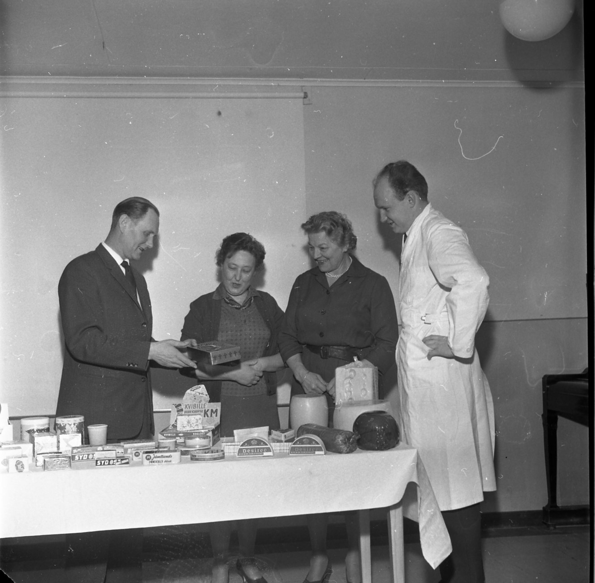 Fyra personer står runt ett bord. En man håller i en ask. På bordet ligger flera olika sorters ostar och en korv.
