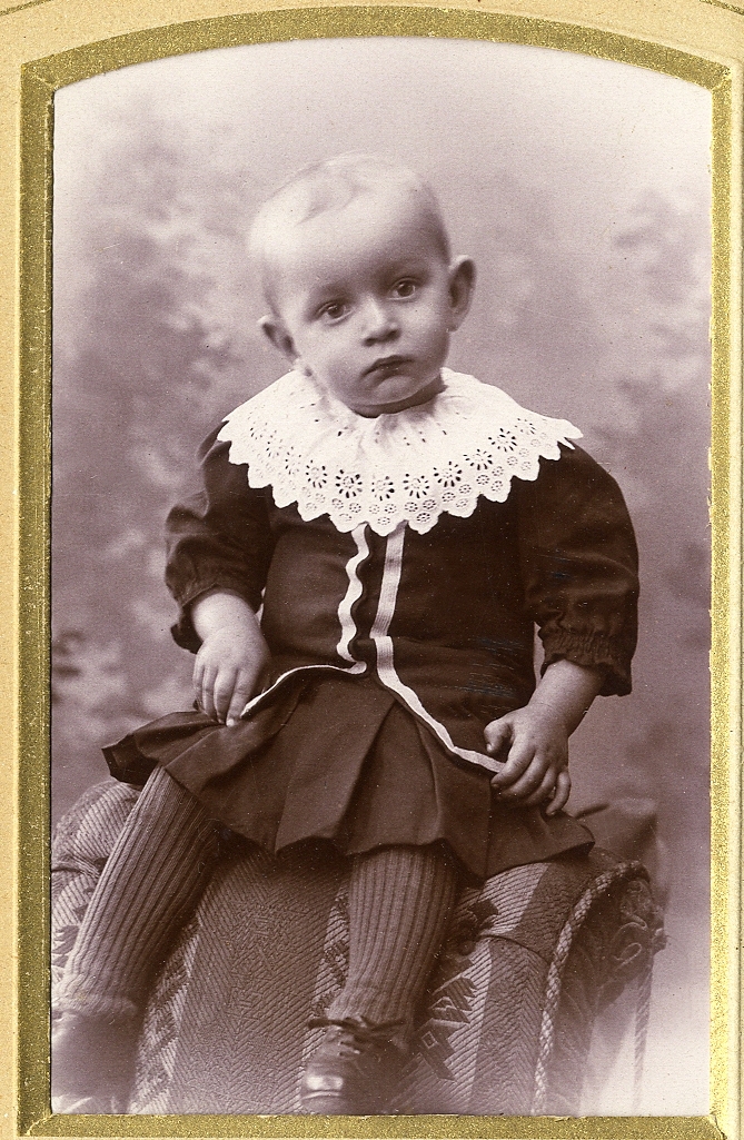 En liten pojke i mörk kolt med stor vit brodyrkrage, sitter på ryggstödet till en stoppad stol.
Helfigur. Ateljéfoto.